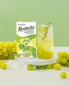 A box of Teazen Kombucha Shina Muscat alongside a glass of the yellow-green kombucha drink, with loose green grapes and a single packet on a white surface against a light green background.