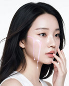 A close-up portrait of a woman with long black hair, wearing a white sleeveless top, with pink cream or serum dripping down her cheek against a clean white background.