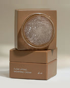 A stacked cosmetic jar set labeled 'FLOW LIFTING WRAPPING CREAM' in a warm taupe-brown color, with the top jar open to reveal a clear gel-like cream inside, on a neutral beige background.
