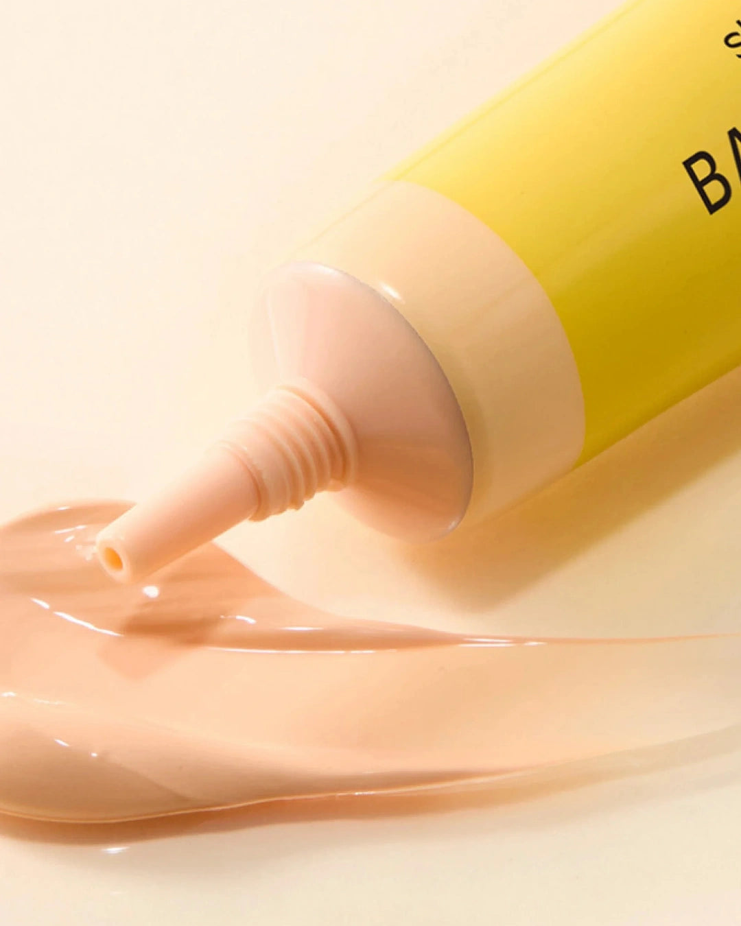 A close-up of a yellow cosmetic tube dispensing a smooth, glossy peach-beige cream onto a light beige surface.