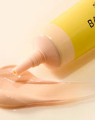 A close-up of a yellow cosmetic tube dispensing a smooth, glossy peach-beige cream onto a light beige surface.