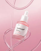Medicube Pion Pink Perfume Serum in a frosted glass dropper bottle with a white label and white dropper cap, set against a soft pink background with abstract curved transparent lines.