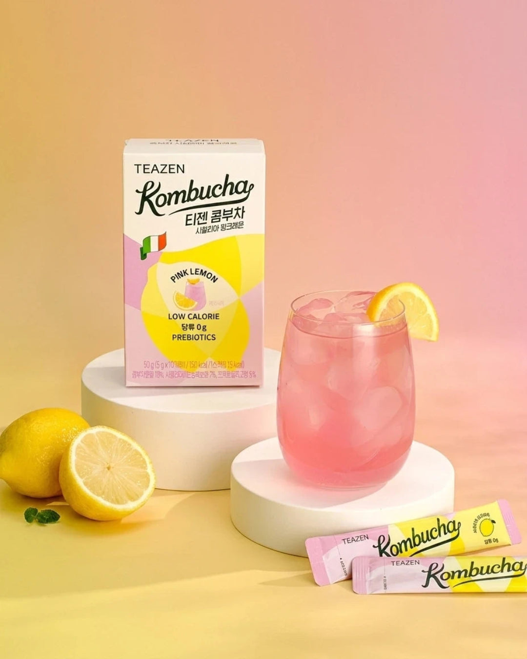A box of TEAZEN Kombucha Pink Lemon flavor is displayed next to a pink drink in a glass with a lemon slice, along with two individual stick packs on a yellow and pink gradient background.