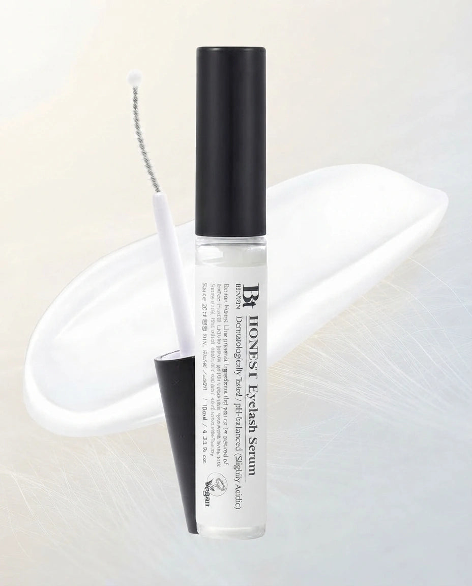HONEST Eyelash Serum in a clear cylindrical bottle with a black cap, shown with the applicator wand and a black cap piece on a soft white background.