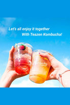 Two hands holding kombucha cups with a blue sky background and text promoting Teazen Kombucha.