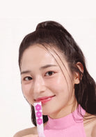 A close-up portrait of a woman with dark hair in a high ponytail, wearing a pink mock-neck top and holding a pink and white flower-patterned stick against her teeth, set against a plain white background.