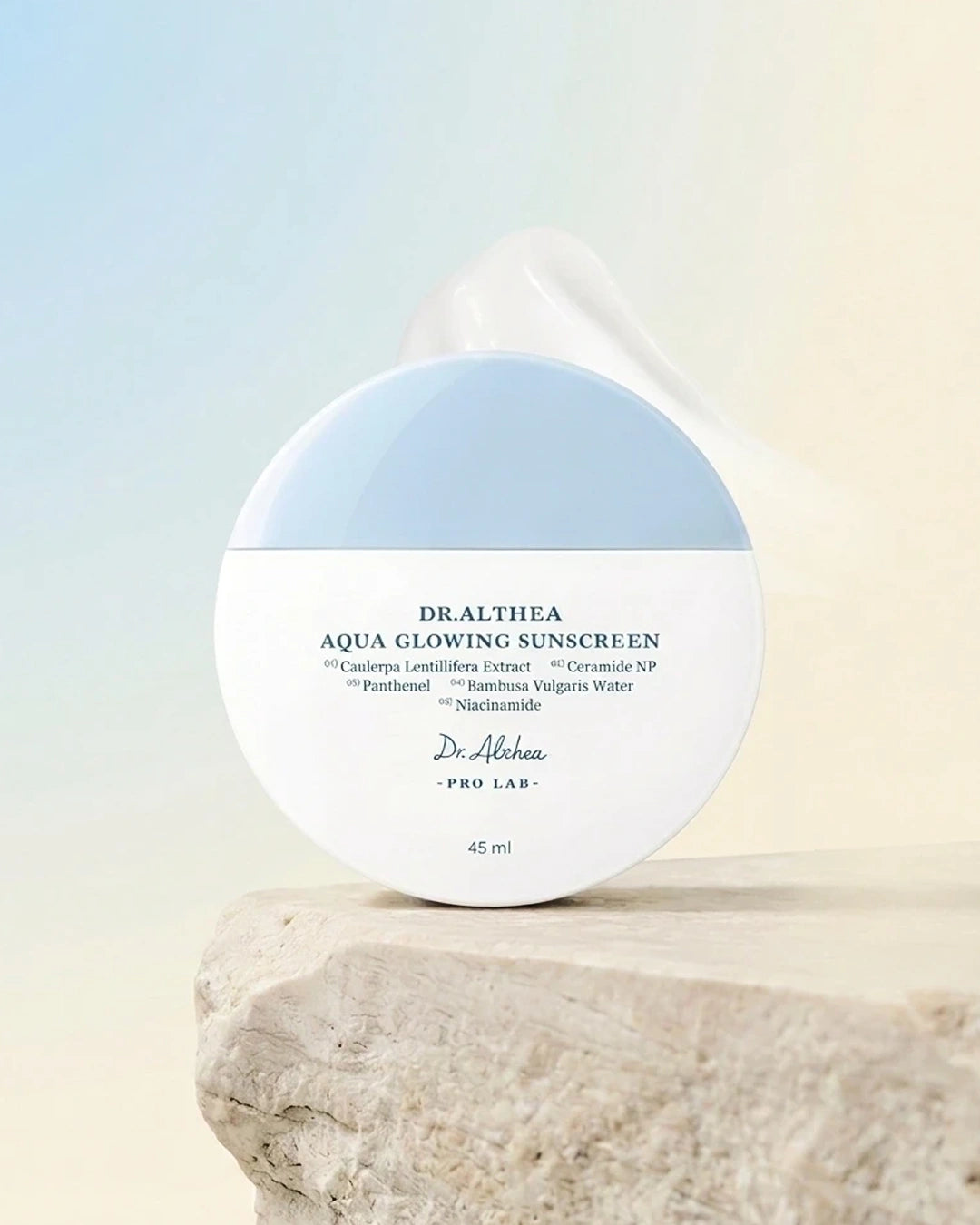 Dr. Alhama Drathea Aqua Glowing Sunscreen in a round white jar with a light blue top section, displayed on a textured stone pedestal against a soft gradient background.